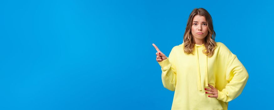 Upset and insecure cute european blond young woman in yellow hoodie, having doubts, being skeptical about topic, pointing finger upper left corner and look unsure camera, blue background.