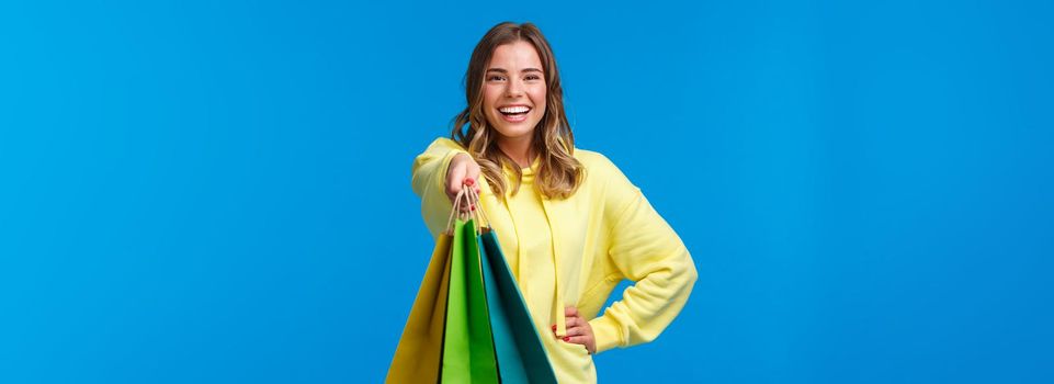 Cheerful blond girl giving you shopping bags and smiling happy, buying presents for family holiday, buying lots of staff in store and asking carry her packages, stand blue background.