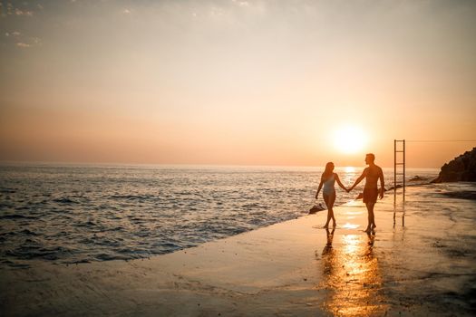 A loving couple walks along the beach by the sea. Young family at sunset by the Mediterranean Sea. Vacation concept. A woman in a swimsuit and a man in shorts at sunset by the sea. Selective focus.