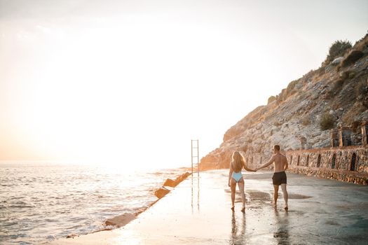 A loving couple walks along the beach by the sea. Young family at sunset by the Mediterranean Sea. Vacation concept. A woman in a swimsuit and a man in shorts at sunset by the sea. Selective focus.