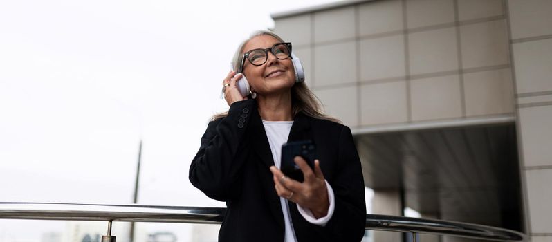 elderly stylish woman listens to music in headphones with a phone in her hands against the backdrop of the building.