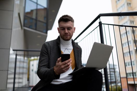 a business man with a laptop and a phone in his hands sits on the stairs next to the office center.