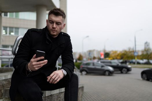 young brutal man in a black coat with a mobile phone on the background of a city parking.
