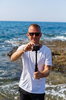 A man in a white T-shirt holds an action camera in his hands and shoots a beautiful view of the sea