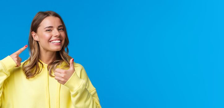 Cheerful and pleased, satisfied good-looking european female pointing at pierced ear and smiling delighted, show thumbs-up like her new jewelry, standing blue background. Copy space
