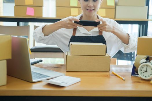 Asian female online business owner or online store owner using cellphone taking picture and checking parcel box, scanning retail package parcel barcode on shipping box preparing for delivery and shipment.