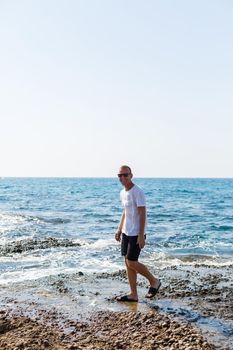 Young attractive man in sunglasses in a white t-shirt and shorts stands on the shore of the mediterranean sea