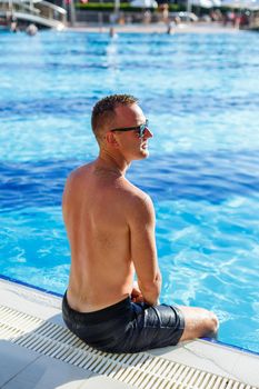 Young attractive man in sunglasses is resting in the pool on summer vacation. The guy in the pool by the hotel