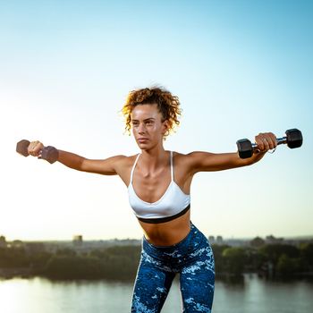 Young fitness woman doing workout with dumbbells by the river in a sunset.