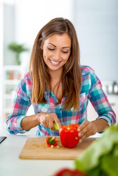 Beautiful young woman making healthy meal in the domestic kitchen. She is cutting red pepper on the kitchen board.