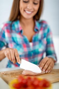 Beautiful young woman making healthy meal in the domestic kitchen. She is cutting chicken meat on the kitchen board. Selective focus. Focus on foreground, on meat.