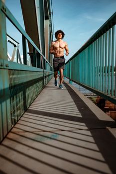 Fit muscular young male runner with naked torso, sprinting at great speed outdoors on the bridge.