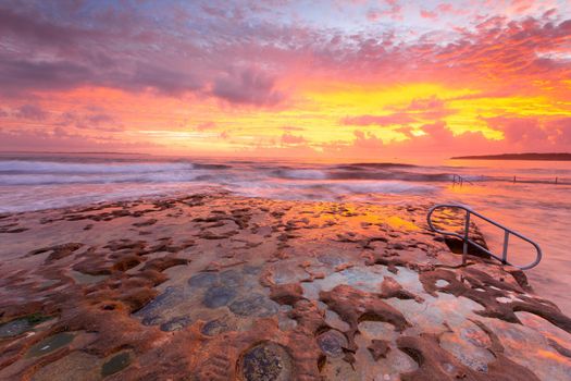 Incoming waves overflow onto the eroded rockshelf and rockpool  with amazing  sunrise sky full of colours