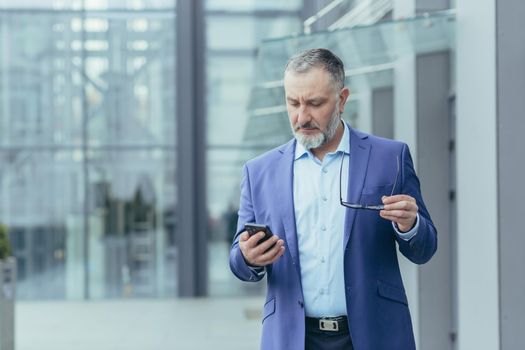 Senior handsome gray-haired man in a suit holding a phone and glasses. Concentratedly reads the news on the phone, looks at messages. It is located near a modern office center.