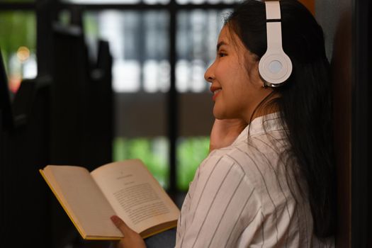 Back view of happy young woman wearing wireless headphone reading book in contemporary library.