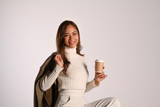 Portrait of modern young woman holding paper cup, sitting on white background. Studio photo, Autumn and Winter concept.