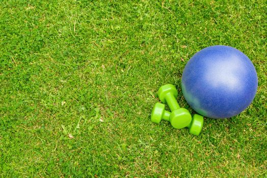 Green ladies dumbbells and blue ball on the green grass background, top view. Outdoor training concept. Copy-space
