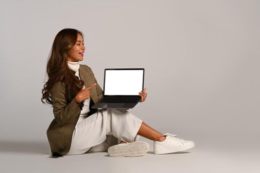 Stylish young woman pointing on laptop screen, sitting on white background. Blank screen for your advertise text.