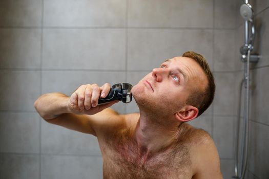 A man shaves his face with an electric razor in front of a mirror. Skin irritation. Bath procedure