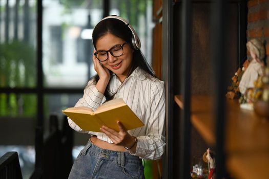 Positive young woman woman wearing headphone and reading book in library. People, knowledge and education concept.