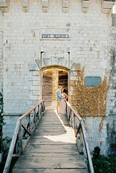 Groom hugs bride leaning against the railing of the wooden bridge to Mamula Fort. High quality photo