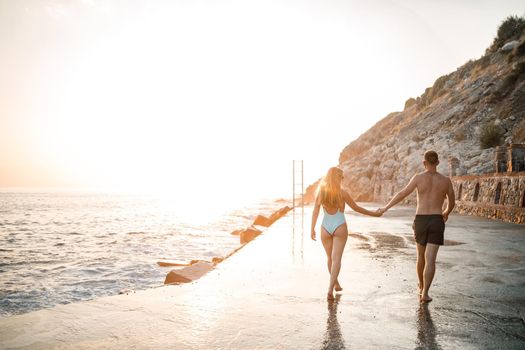 A loving couple walks along the beach by the sea. Young family at sunset by the Mediterranean Sea. Vacation concept. A woman in a swimsuit and a man in shorts at sunset by the sea. Selective focus.