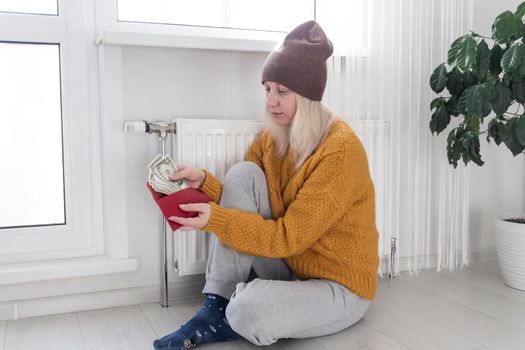 A young girl in a yellow sweater and a brown hat is sitting on the floor, counting money and thinking how to pay bills and taxes near a heater with a thermostat..