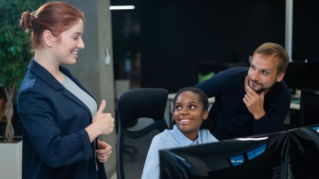 Colleagues look at the monitor and decide working moments. African young woman, caucasian man and red-haired caucasian woman communicate in the office