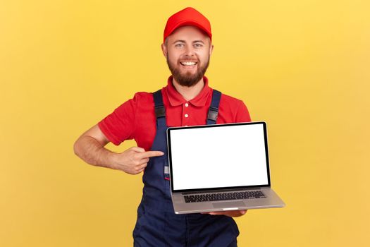 Delighted happy worker man in uniform standing and pointing at laptop blank screen, advertisement area, looking at camera with toothy smile. Indoor studio shot isolated on yellow background.