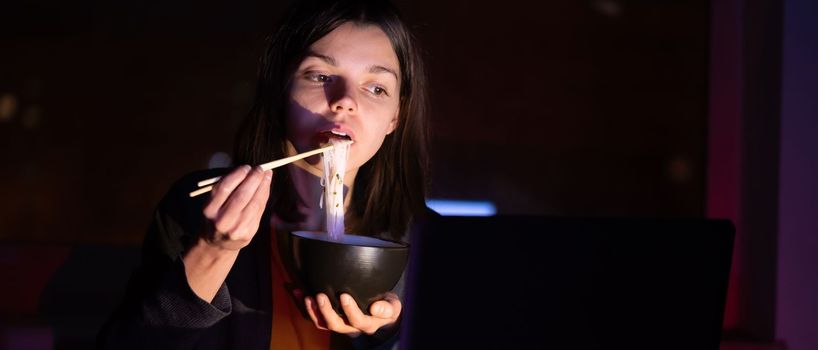 Young girl works at night at home with laptop and eats noodles, asian food with chopsticks. A woman watches a show, a movie and has dinner with ordered food from a cafe.