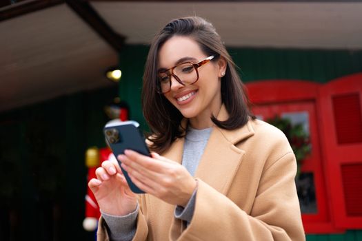 Woman using smartphone on christmas market. Caucasian female enjoying winter holiday season. Concept of social networking, communicating, using app. Looking device screen and smile. Holiday