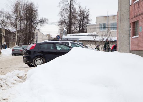 A large snowdrift on the background of a city street. On the road lies white snow in high heaps. Urban winter landscape. Cloudy winter day, soft light.
