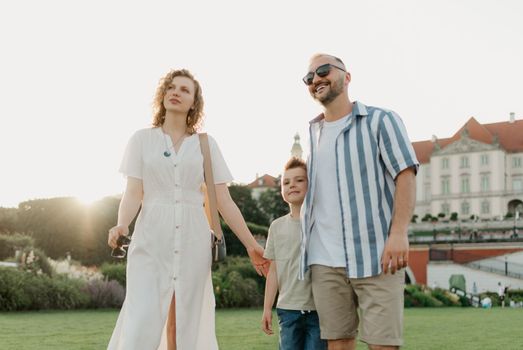 Father, mother and son are hugging in the garden of a European town. Happy family in the evening. Dad is laughing with his family in the background of the palace at sunset.