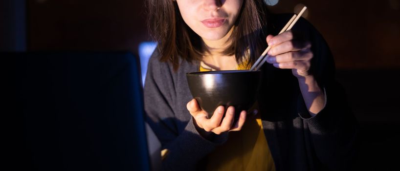 Young girl works at night at home with laptop and eats noodles, asian food with chopsticks. A woman watches a show, a movie and has dinner with ordered food from a cafe.