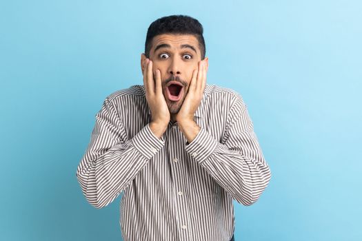 Surprised bearded businessman standing with open mouth and keeps hands on cheek, being impressed of shocking news, wearing striped shirt. Indoor studio shot isolated on blue background.