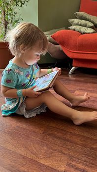 Toddler Girl wearing a turqouise dress sitting on the floor with a Tablet and watching it.
