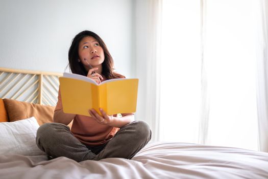 Teen asian girl sitting on bed thinking, using diary and pen to write down thoughts and ideas. Copy space. Lifestyle concept.