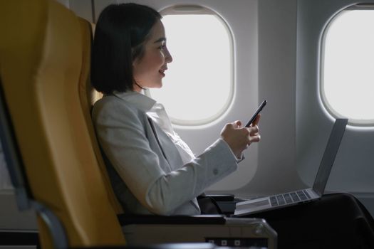 Asian woman passenger sitting in airplane near window and reading news from social networks or using travel applications in smartphone.