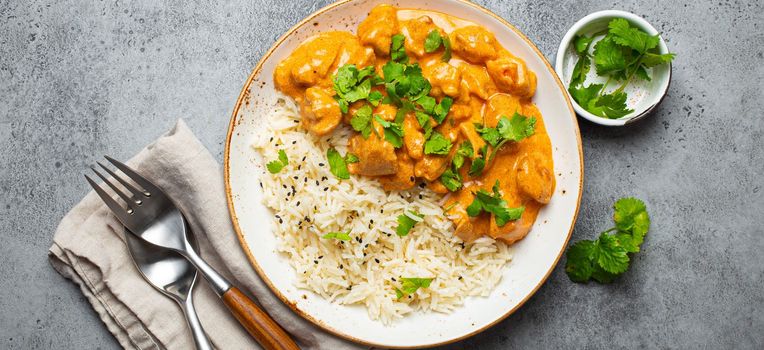 Traditional Indian dish chicken curry with basmati rice and fresh cilantro on rustic white plate on gray concrete table background from above. Indian dinner meal