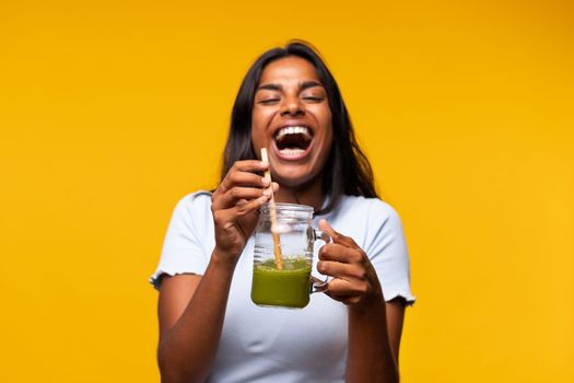 Happy Indian woman with green smoothie on yellow background, laughing. Healthy lifestyle concept.
