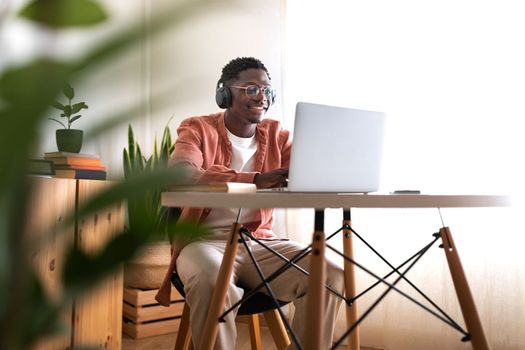 Long shot of young African American man wearing headphones working, studying at home using laptop. Copy space. E-learning and technology concept.