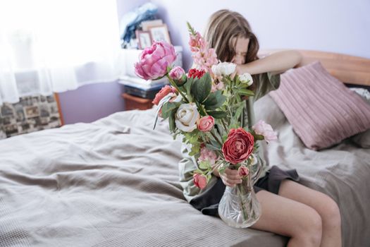 Teenager girl holding a bunch of pink and red peonies in glass vase while closing her face with branches while sitting on sofa. High quality photo
