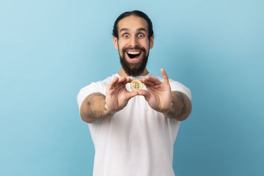 Portrait of surprised man with beard wearing white T-shirt standing holding out bitcoin, paying attention at new digital cryptocurrency. Indoor studio shot isolated on blue background.