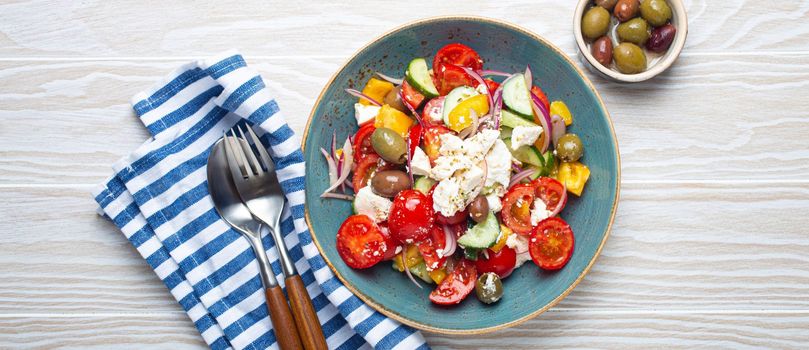 Greek fresh healthy colorful salad with feta cheese, vegetables, olives in blue ceramic bowl on rustic white wooden background top view, Mediterranean diet, traditional cuisine of Greece