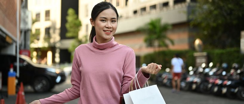 Smiling asian woman enjoying shopping, carrying shopping bags walking in city streets on sunny day.