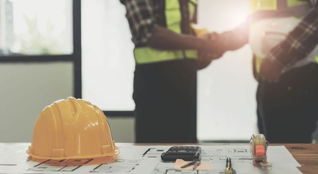 yellow safety helmet on workplace desk with construction worker team hands shaking greeting start up plan new project contract in office center at construction site, partnership and contractor concept..
