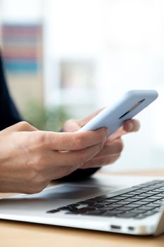 Vertical close-up image of woman hands using mobile phone while working in the office with laptop. Selective focus on hands. Blurred background. Technology concept.