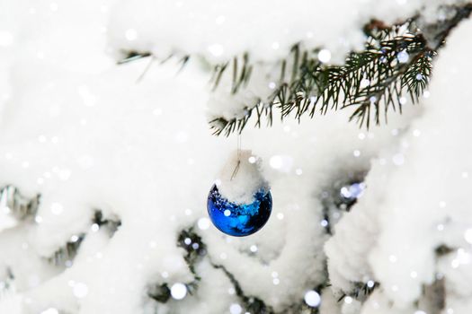 A branch of a Christmas tree in the snow with a Christmas decoration. A blue Christmas toy is hanging on a tree outside. Snowfall. Lots of snow. Winter, festive background.