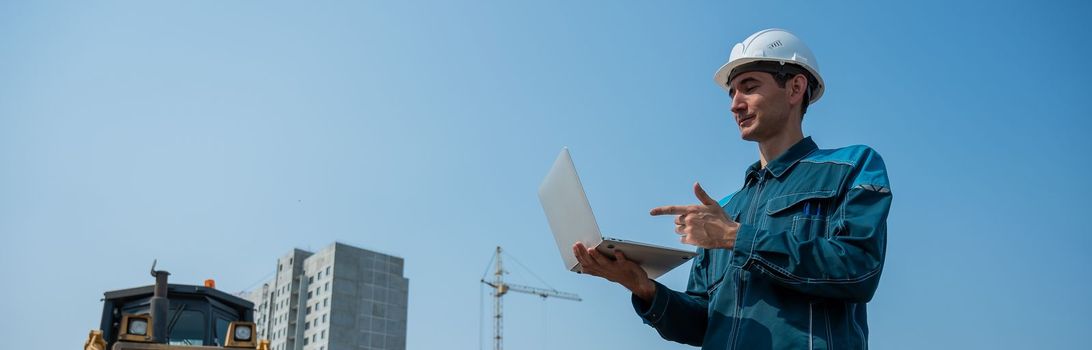 A male builder in a helmet stands against the background of a construction site with a laptop in his hands. Widescreen