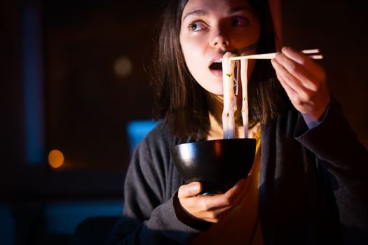 Young girl works at night at home with laptop and eats noodles, asian food with chopsticks. A woman watches a show, a movie and has dinner with ordered food from a cafe.